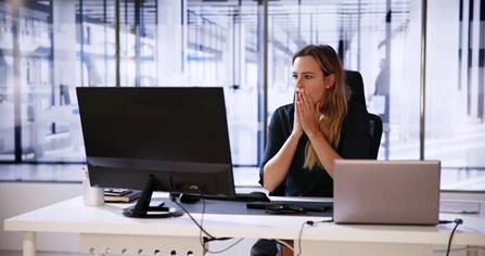 A worried woman sits at her desk with a computer and laptop.